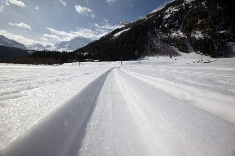 Gespurte Langlaufloipe im verschneiten Gebirgstal unter blauem Himmel mit umliegenden Wäldern und Berggipfeln.