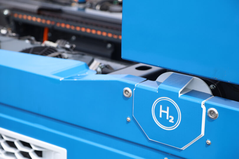 Nahaufnahme eines blauen Wasserstofffahrzeugs mit dem Symbol „H₂“ auf der Abdeckung. Im Hintergrund sind technische Komponenten und Kabel sichtbar, die auf ein modernes Antriebssystem hinweisen.
