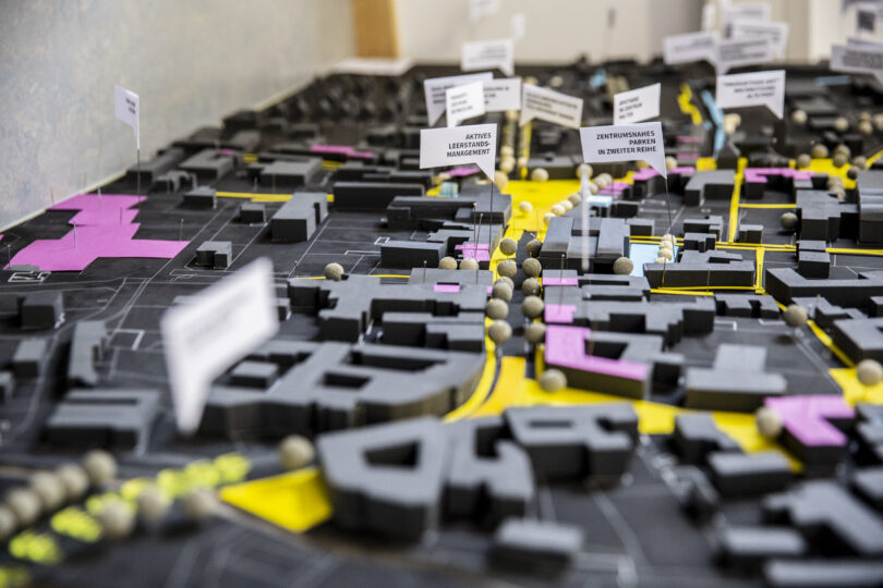 Städtebauliches Modell mit Gebäuden, Straßen und beschrifteten Markierungen. Gelbe und pinke Flächen heben bestimmte Zonen hervor, kleine Schilder kennzeichnen Planungsmaßnahmen wie „Leerstandsmanagement“ oder „zentrumsnahes Parken“.