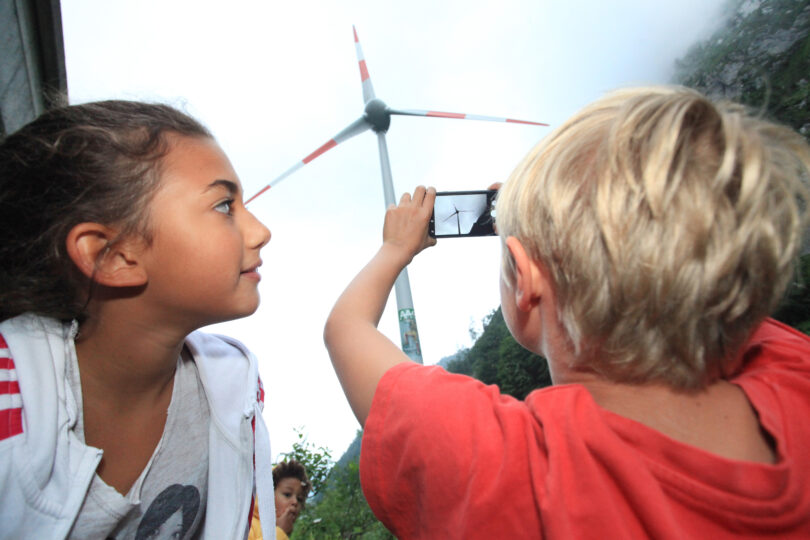 Zwei Kinder betrachten eine große Windkraftanlage im Hintergrund. Eines der Kinder hält ein Smartphone und fotografiert das Windrad. Im Hintergrund sind Nebel und bewaldete Hänge zu sehen.