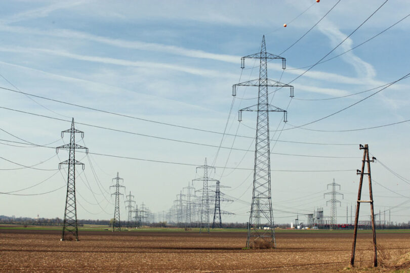 : Mehrere Hochspannungsmasten verlaufen über ein weites, braunes Feld. Die Stromleitungen spannen sich in Reihen bis zum Horizont, darüber ein klarer Himmel mit leichten Wolkenstreifen.