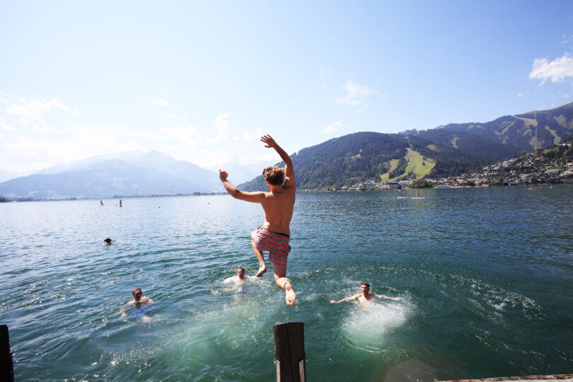 Mehrere Menschen baden in einem klaren See, während eine Person in bunter Badehose gerade von einem Steg ins Wasser springt. Im Hintergrund sind Berge, Wälder und Häuser unter blauem Himmel zu sehen.