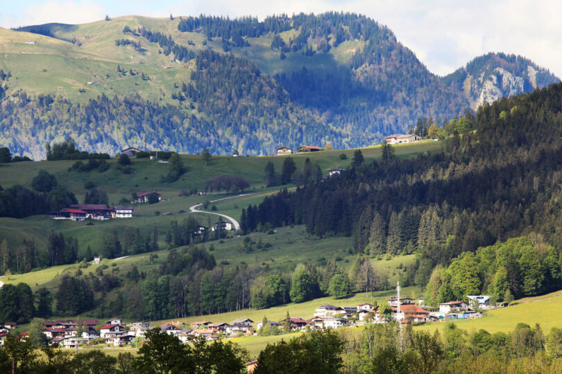 Grünes Alpendorf in hügeliger Landschaft mit verstreuten Bauernhöfen und Wiesen. Im Hintergrund erheben sich bewaldete Berge, die teils im Schatten liegen, unter einem leicht bewölkten Himmel.