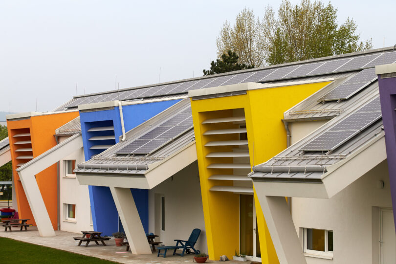 Reihenhausanlage mit bunten Fassaden in Orange, Blau, Gelb und Violett. Auf den schrägen Dächern sind Solarpaneele installiert. Vor den Häusern stehen Sitzbänke und Pflanzen, im Hintergrund Bäume.