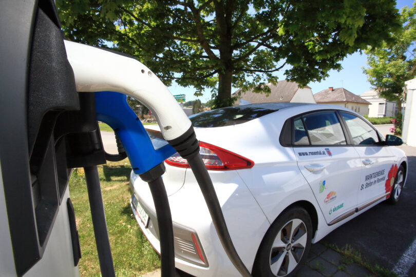 Ein weißes Elektroauto ist an einer Ladesäule angeschlossen, während es im Schatten eines Baums steht. Im Vordergrund sind zwei Ladestecker zu sehen, einer davon verbunden mit dem Fahrzeug.
