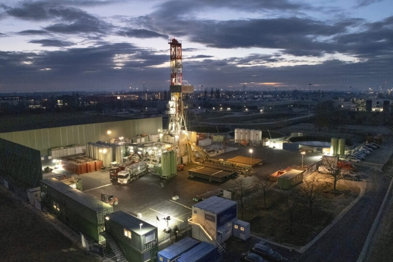 Beleuchtete Geothermie-Bohranlage in der Dämmerung. Auf dem Gelände sind Bohrtürme, Container, Fahrzeuge und technische Geräte zu sehen. Im Hintergrund leuchten Stadtlichter unter einem bewölkten Abendhimmel.