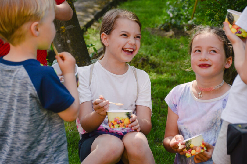 Mehrere Kinder sitzen im Garten auf dem Rasen, essen Eis aus bunten Bechern und lachen fröhlich. Sonnenlicht fällt durch die Bäume und sorgt für eine sommerlich fröhliche Atmosphäre.