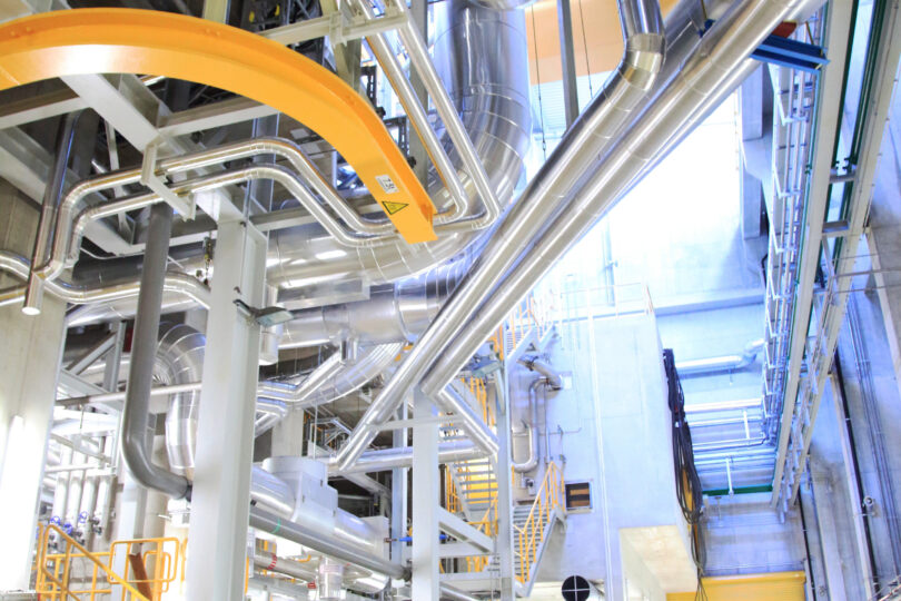 Innenansicht einer modernen Industrieanlage mit großen Edelstahlrohren, Wartungsstegen und einer Person mit Warnweste und Helm auf einer Begehung.