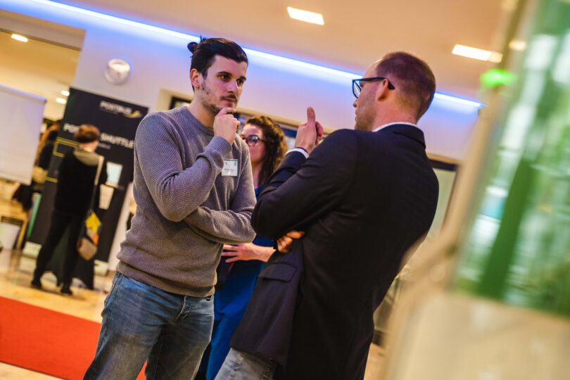 Zwei Männer führen ein Gespräch in einem modernen Innenraum mit Konferenzatmosphäre. Einer trägt einen Anzug, der andere Freizeitkleidung. Im Hintergrund sind weitere Personen und Infostände zu sehen.