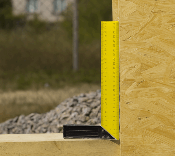 Gelber Winkelmesser aus Metall misst den rechten Winkel zwischen einer Holzplatte und einem horizontalen Brett auf einer Baustelle im Freien.