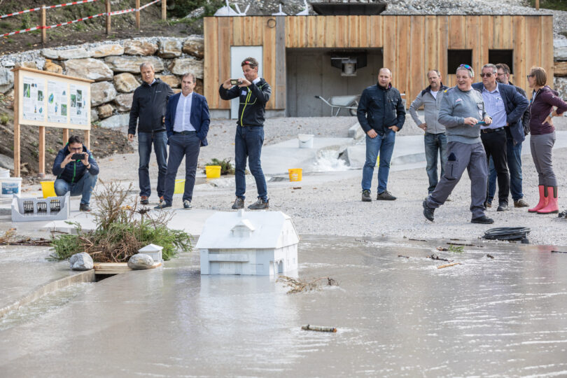 Gruppe von Personen beobachtet ein Modellversuchsgelände mit Wasserlauf und Miniaturhäusern. Das Szenario simuliert Überflutung oder Hochwasser, während Teilnehmende Fotos machen und sich austauschen.