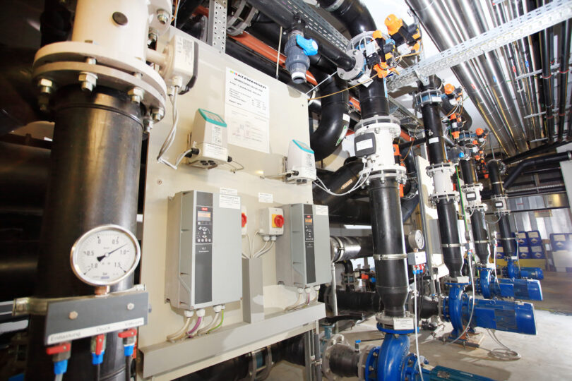 Technikraum mit Rohrleitungen, Pumpen und Messinstrumenten einer Energie- oder Wasserversorgungsanlage. Sichtbar sind Druckanzeigen, Steuergeräte und Ventile, die den technischen Betrieb überwachen.