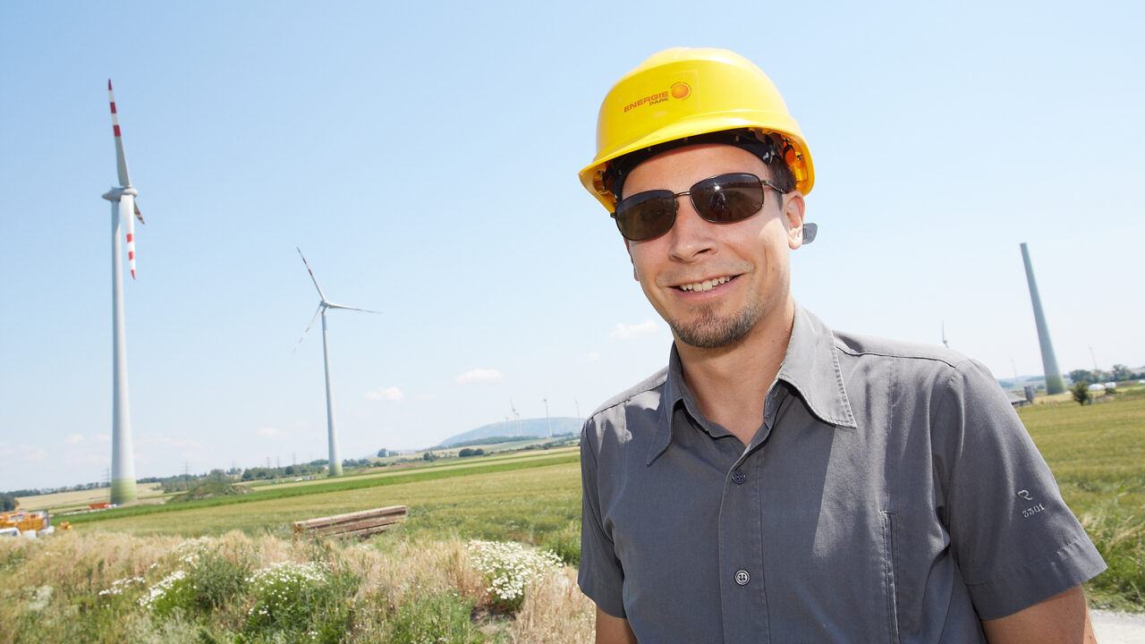 Ein Mann mit Schutzhelm und Sonnenbrille steht auf einem Windparkgelände. Hinter ihm sind mehrere Windräder auf grünen Feldern zu sehen, der Himmel ist klar und sonnig.
