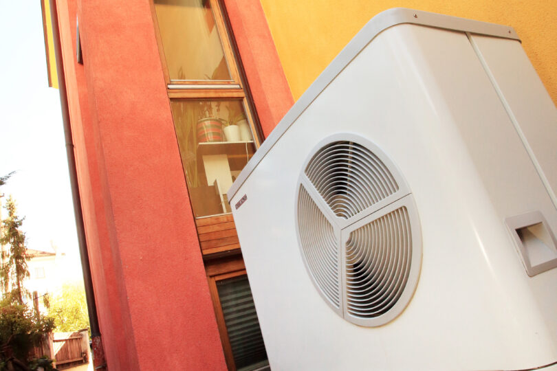 Wärmepumpe neben einem modernen Wohngebäude mit farbigen Fassaden in Rot und Gelb. Das Gerät steht auf einem gepflasterten Weg, im Hintergrund sind Fenster und Pflanzen zu sehen.