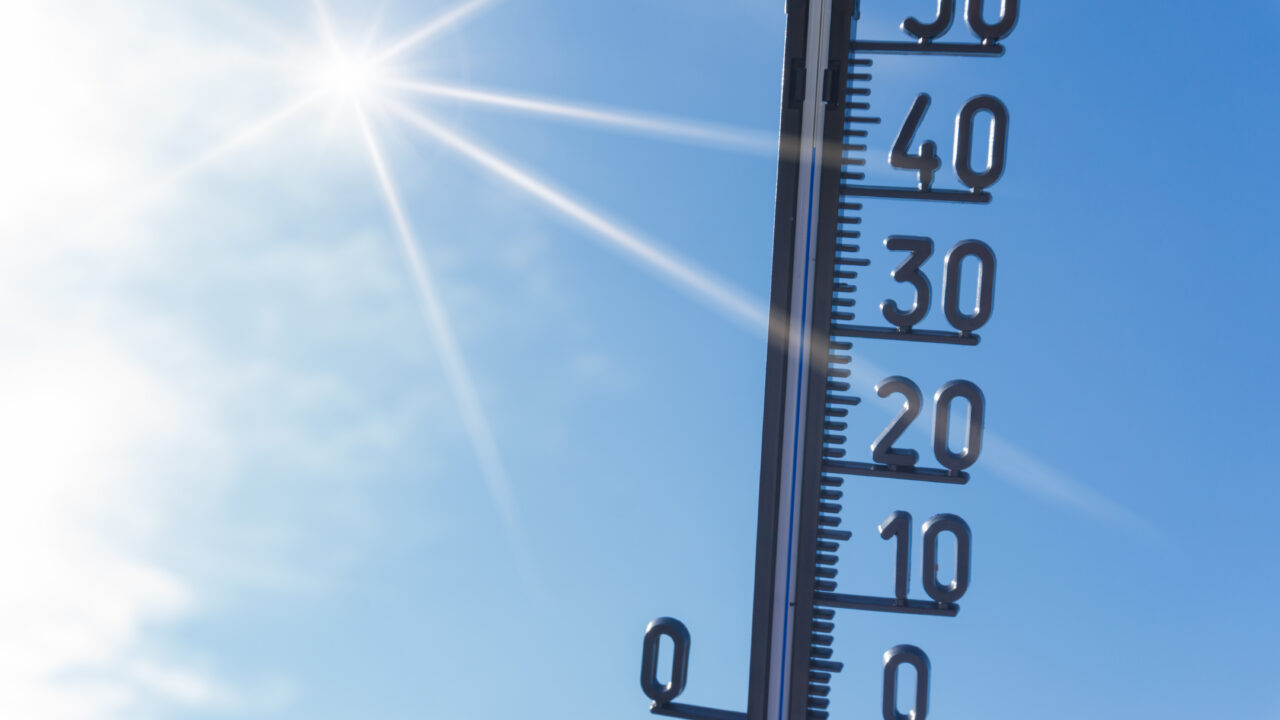 Im Hintergrund sieht man den blauen, wolkenlosen Himmel. Links oben scheint die Sonne. Es wird ein Thermometer hochgehalten.