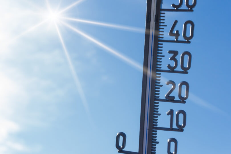 Im Hintergrund sieht man den blauen, wolkenlosen Himmel. Links oben scheint die Sonne. Es wird ein Thermometer hochgehalten.