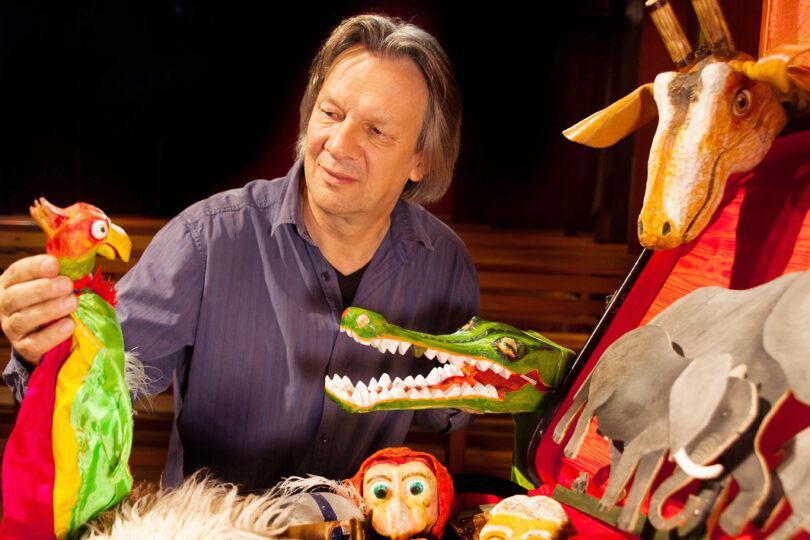 Ein Mann steht auf einer Bühne und hält bunte Handpuppen in der Hand. Vor ihm liegen zahlreiche Figuren und Tierpuppen, darunter ein Krokodil, ein Papagei und eine Giraffe. Die Szene ist warm beleuchtet.