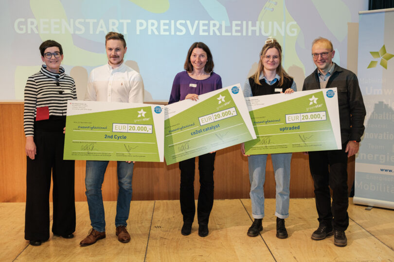 Drei Gewinnerteams der Greenstart-Preisverleihung halten symbolische Schecks über je 20.000 Euro auf einer Bühne, flankiert von zwei Vertreter:innen der Veranstalter.