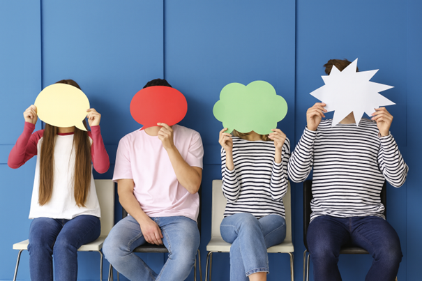 Vier Personen sitzen nebeneinander auf Stühlen und halten bunte Sprechblasen-Schilder vor ihre Gesichter vor blauem Hintergrund.