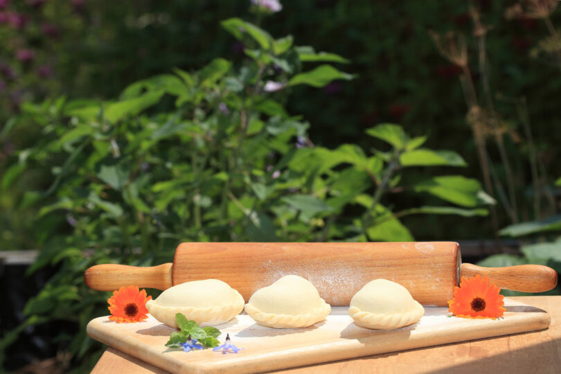 Drei ungekochte Teigtaschen liegen auf einem Holzbrett mit Nudelholz, verziert mit essbaren Blüten und Kräutern. Im Hintergrund ist grüne Vegetation zu sehen, das Bild wirkt sommerlich und natürlich.