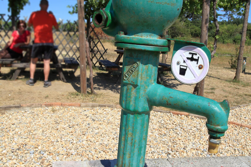 Grüne historische Wasserpumpe mit Trinkwasser-Verboten-Symbol auf einem gemauerten Sockel, daneben ein Zollstock. Im Hintergrund sitzen Menschen unter einer Pergola mit Weinreben an einem sonnigen Tag. Symbolisiert Hitze und Wasserknappheit.