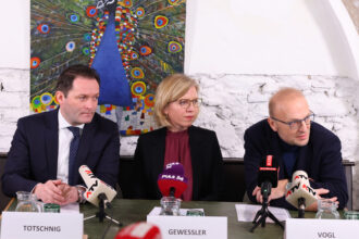Drei Personen sitzen nebeneinander bei einer Pressekonferenz an einem Tisch. Vor ihnen stehen Mikrofone verschiedener Medien, darunter ATV, PULS 24 und KURIER. Namensschilder mit den Aufschriften „TOTSCHNIG“, „GEWESSLER“ und „VOGL“ markieren ihre Plätze. Im Hintergrund ist ein buntes Gemälde eines Pfaus zu sehen, das die Szene lebhaft ergänzt. Die Personen wirken konzentriert und aufmerksam, während einer von ihnen spricht.