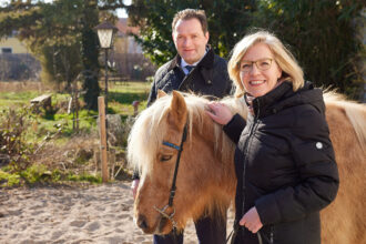 Eine Frau und ein Mann stehen im Freien neben einem braunen Pony mit heller Mähne. Die Frau im Vordergrund lächelt freundlich in die Kamera, während sie das Pony streichelt. Sie trägt eine schwarze Winterjacke und eine Brille. Der Mann im Hintergrund, ebenfalls in einer dunklen Jacke, schaut ebenfalls in die Kamera. Im Hintergrund ist ein Garten mit Bäumen, Sträuchern und einer alten Laterne zu sehen. Die Szene wirkt sonnig und freundlich.