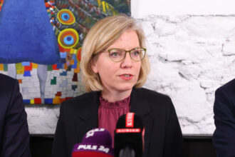 Eine Frau mit blonden, kinnlangen Haaren und einer grünen Brille spricht bei einer Pressekonferenz. Sie trägt eine schwarze Blazerjacke und eine bordeauxfarbene Bluse. Vor ihr stehen mehrere Mikrofone verschiedener Medien, darunter „PULS 24“ und „KURIER“. Im Hintergrund ist eine weiße Backsteinwand und ein farbenfrohes Gemälde eines Pfaus zu sehen.