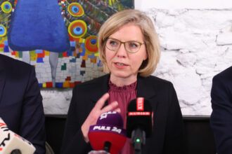 Eine Frau mit blonden, kinnlangen Haaren und einer grünen Brille spricht bei einer Pressekonferenz. Sie trägt eine schwarze Blazerjacke und eine bordeauxfarbene Bluse. Vor ihr stehen mehrere Mikrofone verschiedener Medien, darunter „PULS 24“ und „KURIER“. Im Hintergrund ist eine weiße Backsteinwand und ein farbenfrohes Gemälde eines Pfaus zu sehen.