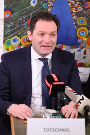Ein Mann in einem dunkelblauen Anzug mit weißem Hemd und dunkelblauer Krawatte mit Punkten spricht bei einer Pressekonferenz. Vor ihm stehen mehrere Mikrofone, darunter eines vom „KURIER“ und ein weiteres von „ATV“. Auf dem Tisch liegt ein Namensschild mit der Aufschrift „TOTSCHNIG“. Im Hintergrund ist ein buntes Gemälde eines Pfaus mit farbenfrohen Augenfedern zu sehen.