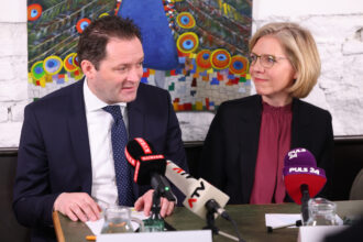 Ein Mann im dunkelblauen Anzug mit weißem Hemd und blauer Krawatte mit Punkten spricht bei einer Pressekonferenz, während eine Frau mit blonden Haaren, Brille und bordeauxroter Bluse neben ihm sitzt und ihn ansieht. Vor ihnen stehen Mikrofone von „KURIER“, „ATV“ und „PULS 24“. Im Hintergrund ist ein buntes Pfauen-Gemälde zu sehen.