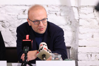 Ein Mann mit Glatze, Brille mit blauen Rahmen und dunklem Sakko spricht bei einer Pressekonferenz. Vor ihm stehen Mikrofone von „KURIER“ und „ATV“. Im Hintergrund ist eine weiße, gemauerte Wand zu sehen. Der Mann wirkt konzentriert und engagiert im Gespräch.