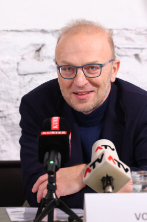 Ein Mann mit Glatze, Brille mit blauen Rahmen und dunklem Sakko spricht bei einer Pressekonferenz. Vor ihm stehen Mikrofone von „KURIER“ und „ATV“. Im Hintergrund ist eine weiße, gemauerte Wand zu sehen. Der Mann wirkt konzentriert und engagiert im Gespräch.