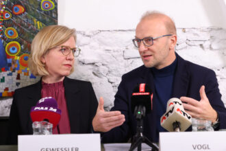 Zwei Personen, eine Frau mit blonden Haaren und Brille (GEWESSLER) sowie ein Mann mit Brille und Glatze (VOGL), sitzen nebeneinander bei einer Pressekonferenz. Vor ihnen stehen Mikrofone von „PULS 24“, „KURIER“ und „ATV“. Der Mann gestikuliert mit den Händen, während die Frau ihn aufmerksam ansieht. Im Hintergrund ist eine weiße Backsteinwand mit einem bunten Gemälde eines Pfaus zu sehen.