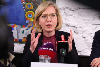 Eine Frau mit blonden, schulterlangen Haaren, einer Brille mit grünlichem Rahmen und einem dunklen Blazer über einer bordeauxfarbenen Bluse spricht auf einer Pressekonferenz. Sie gestikuliert mit beiden Händen, als wolle sie etwas betonen. Vor ihr stehen mehrere Mikrofone von Medien wie „PULS 24“ und „KURIER“. Im Hintergrund ist eine weiße Ziegelwand und ein buntes Gemälde mit einem stilisierten Pfau zu sehen.