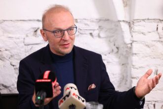 Ein Mann mit Glatze, Brille mit blauen Rahmen und dunklem Sakko spricht bei einer Pressekonferenz. Vor ihm stehen Mikrofone von „KURIER“ und „ATV“. Im Hintergrund ist eine weiße, gemauerte Wand zu sehen. Der Mann wirkt konzentriert und engagiert im Gespräch.