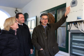 Drei Personen stehen neben einer weißen Wand mit einem grünen Fensterrahmen. Ein Mann in einer dunklen Jacke zeigt auf ein technisches Gerät mit mehreren Kabeln, vermutlich ein Steuerungspanel oder ein Wechselrichter für Solaranlagen. Er erklärt offensichtlich etwas den beiden anderen Personen, einer Frau mit blonden Haaren und Brille sowie einem Mann im Anzug mit gestepptem Mantel. Beide hören aufmerksam zu und lächeln. Im Hintergrund hängt ein dekorativer Kranz an der Wand. Die Szene wirkt wie eine Besichtigung oder technische Präsentation.