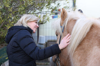 Eine Frau mit blonden Haaren und Brille, gekleidet in eine schwarze Winterjacke, streichelt sanft den Hals eines braunen Ponys mit heller Mähne. Beide wirken entspannt und zufrieden. Im Hintergrund sind grüne Pflanzen und ein Zaun zu sehen, was darauf hindeutet, dass sich die Szene im Freien abspielt.