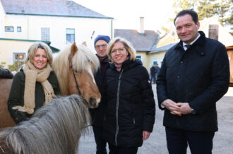 Vier Personen, zwei Frauen und zwei Männer, stehen draußen vor einem Gebäude mit gelber Fassade und grünen Fensterläden. Sie tragen warme Winterkleidung, darunter dicke Jacken und Schals. Die Frau links hält ein braunes Pony mit heller Mähne am Zügel, während ein kleineres, graues Pony mit langer Mähne im Vordergrund steht. Alle lächeln in die Kamera, was eine freundliche und entspannte Atmosphäre vermittelt. Im Hintergrund sind weitere Gebäude mit Ziegeldächern und einige Bäume zu sehen.