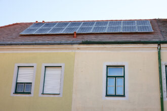 Ein zweistöckiges Gebäude mit gelb-beiger Fassade und grünen Fensterrahmen. Auf dem rot gedeckten Dach sind mehrere Solarpaneele installiert, die zur nachhaltigen Energiegewinnung beitragen. Die Hauswand zeigt leichte Abnutzungsspuren, während ein grünes Fallrohr an der Seite verläuft.