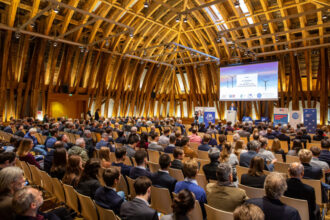 Ein großer Konferenzsaal mit beeindruckender Holzarchitektur ist gefüllt mit Zuhörern, die einem Vortrag lauschen. Auf der Bühne steht ein Redner am Podium, während eine große Leinwand hinter ihm die Präsentation „The Highlights of the World Energy Outlook 2022“ zeigt. Verschiedene Banner, unter anderem von der TU Wien und dem World Energy Council, flankieren die Bühne.
