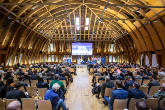 Ein weitläufiger Konferenzsaal mit beeindruckender Holzarchitektur, gefüllt mit zahlreichen Teilnehmern, die einer Präsentation folgen. Auf der großen Leinwand ist eine Folie mit Windkraftanlagen und dem Titel „The Highlights of the World Energy Outlook 2022“ zu sehen. Vorne auf der Bühne stehen mehrere Redner, während das Publikum in Reihen von Stühlen konzentriert zuhört.
