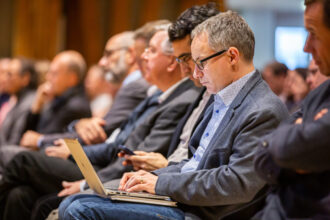 Ein Mann mit Brille sitzt in einem Konferenzsaal und arbeitet konzentriert an seinem Laptop, während er an einer Veranstaltung teilnimmt. Neben ihm nutzen andere Teilnehmer ihre Smartphones oder hören der Präsentation zu. Der Raum ist mit Menschen gefüllt, die in formeller Kleidung an der Konferenz teilnehmen.