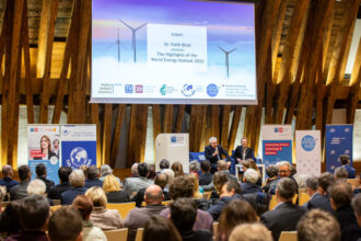 Ein Konferenzsaal mit Holzverkleidung ist gefüllt mit Publikum, das einer Podiumsdiskussion lauscht. Auf der Bühne sitzen zwei Personen vor einer großen Leinwand, auf der eine Präsentation mit dem Titel „The Highlights of the World Energy Outlook 2022“ angezeigt wird. Verschiedene Banner im Raum zeigen die Logos der TU Wien, des Klima- und Energiefonds sowie des World Energy Councils.