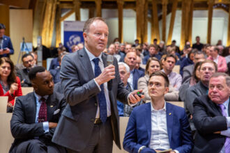 Ein Mann im Anzug steht in einem gut gefüllten Konferenzsaal und hält ein Mikrofon, während er eine Frage stellt oder eine Aussage trifft. In seiner anderen Hand hält er einen Präsentationsklicker. Das Publikum im Hintergrund hört aufmerksam zu, einige Personen sitzen mit verschränkten Armen oder nehmen Notizen. Der Raum hat eine warme Holzarchitektur, die eine professionelle, aber einladende Atmosphäre schafft.