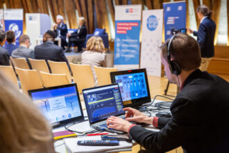 Ein Techniker mit Kopfhörern sitzt an einem Tisch mit mehreren Laptops und einem Mischpult und steuert die audiovisuelle Übertragung einer Konferenz. Im Hintergrund ist eine Bühne mit zwei sprechenden Personen sowie Publikum in einem Saal mit Holzbalken zu sehen. Verschiedene Banner mit Logos und Texten deuten auf eine wissenschaftliche oder wirtschaftliche Veranstaltung hin.