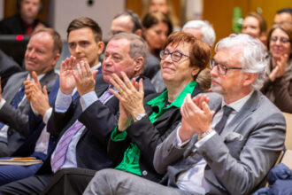 Mehrere Personen in eleganter Kleidung sitzen in einem Konferenzsaal und applaudieren. Eine Frau mit grüner Bluse und Brille lächelt dabei, während ein Mann im grauen Anzug konzentriert klatscht. Im Hintergrund sind weitere Teilnehmerinnen und Teilnehmer sichtbar, die ebenfalls Beifall spenden, was auf einen erfolgreichen Vortrag oder eine besondere Ankündigung hinweist.