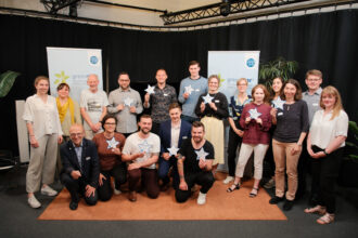 Eine Gruppe von etwa 16 Personen posiert für ein Gruppenfoto vor zwei „greenstart“-Bannern in einem Veranstaltungssaal mit schwarzem Vorhang. Mehrere Personen halten blaue sternförmige Auszeichnungen mit Namen ihrer Projekte, während andere lächeln oder sich leicht zur Kamera neigen. Die Gruppe ist vielfältig, mit unterschiedlichen Altersgruppen und Stilen, einige stehen, während andere knien oder in der Hocke sitzen.