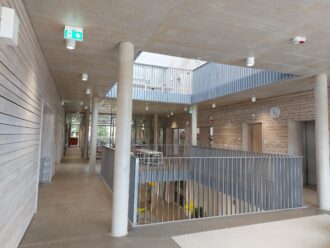 Ein heller, moderner Innenraum eines Schul- oder Bildungsgebäudes mit offenem Treppenhaus. Die Wände sind mit hellem Holz verkleidet, während der Boden aus glattem Beton besteht. Weiße runde Deckenspots sorgen für Beleuchtung. Das Geländer des Treppenhauses und des offenen Flurs im Obergeschoss besteht aus schlichten grauen Metallstreben. An den Wänden befinden sich grüne Notausgangsschilder mit Pfeilen nach unten. Im Hintergrund sind Glaswände zu weiteren Räumen erkennbar, die Tageslicht in das Gebäude lassen. Eine analoge Wanduhr zeigt die Zeit an, und ein Aufzug ist auf der rechten Seite zu sehen. Die Architektur wirkt minimalistisch, offen und funktional.