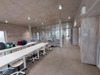 Ein moderner, offener Lernraum mit langen weißen Tischen und höhenverstellbaren Hockern auf Rollen. Auf den Tischen liegen mehrere bunte Schulrucksäcke. In der Mitte des Raumes stehen mobile Bücherregale mit Kinderbüchern und Spielen. Die Wände sind mit hellem Holz verkleidet, die Decke besteht aus Beton mit runden Deckenspots. Durch große Glaswände ist ein weiterer Bereich sichtbar, der ebenfalls als Lern- oder Aufenthaltsraum dient. Der Boden ist aus glattem Beton und vermittelt eine minimalistische, moderne Atmosphäre.